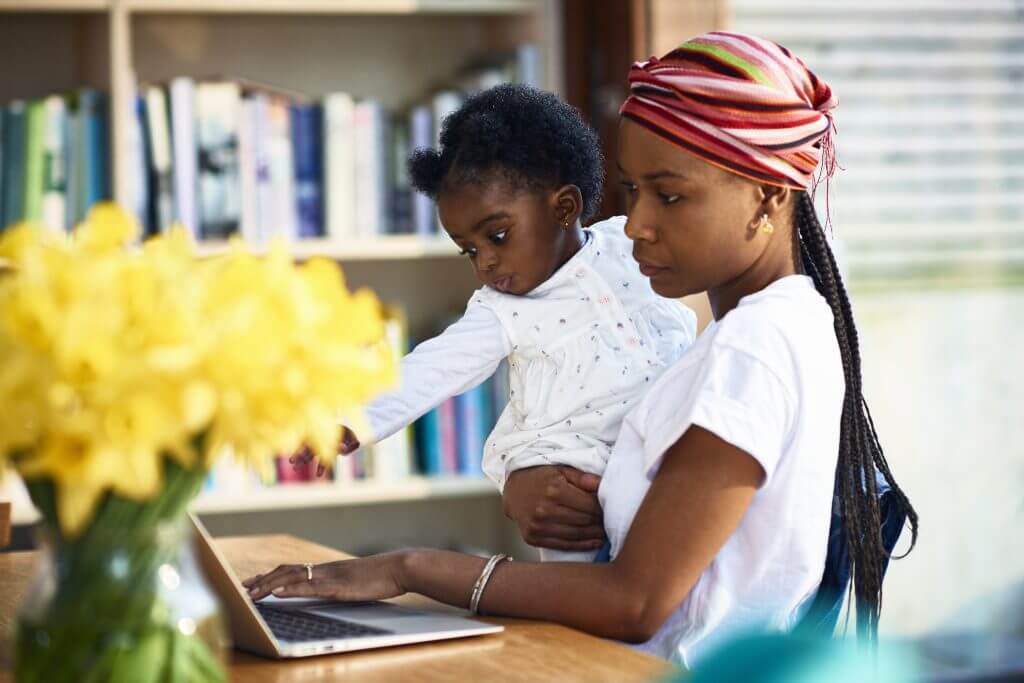 Zwarte vrouw met dochter op de arm surft rechtopstaand op haar laptop.